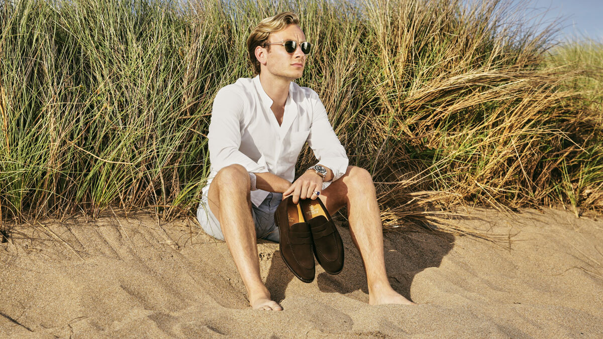 Man in shorts sitting on beach holding pair of suede loafers