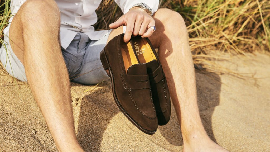 Man wearing shorts and holding suede loafers on the beach